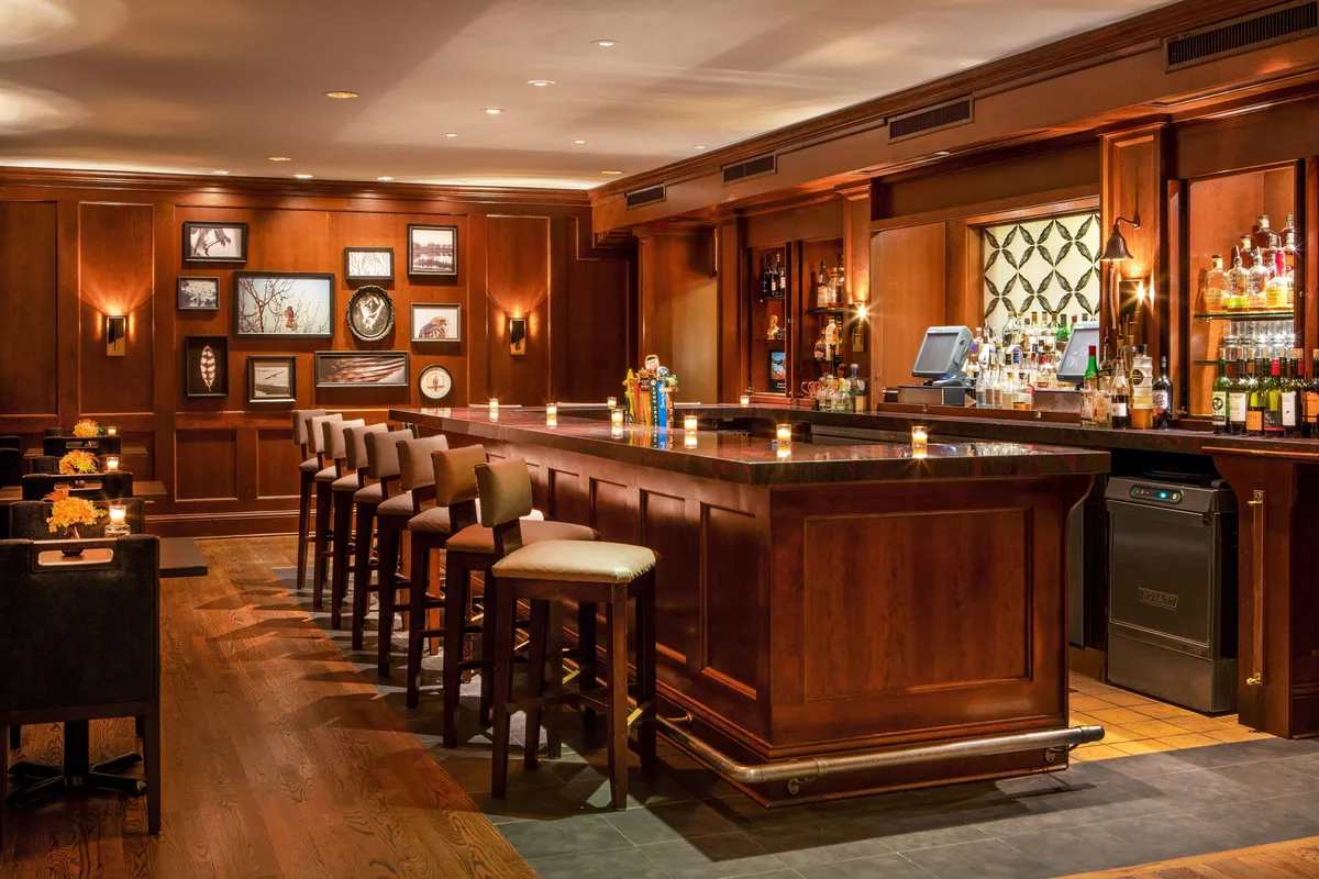 This image showcases an inviting interior bar scene, with a sleek bar counter lined with comfortable seating, bottles of premium spirits artfully displayed on shelves, and tasteful artwork adorning the walls. The ambient lighting and arrangement encourage guests to gather for drinks, engage in intimate conversations, and celebrate special moments in a cozy yet sophisticated setting.