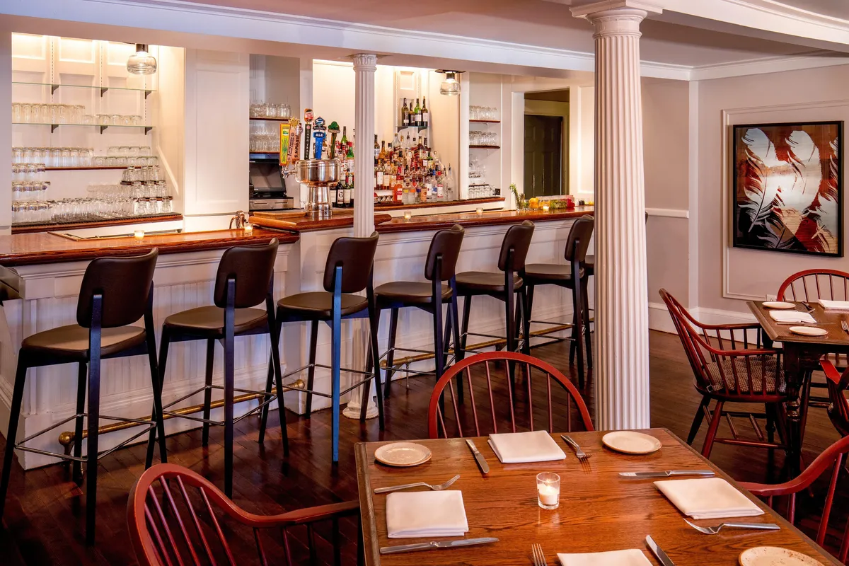 A vibrant bar and dining area featuring wooden tables, bar stools, and colorful artwork on the walls. The scene captures guests enjoying drinks, dining, and lively discussions, illustrating an energetic and welcoming communal space.