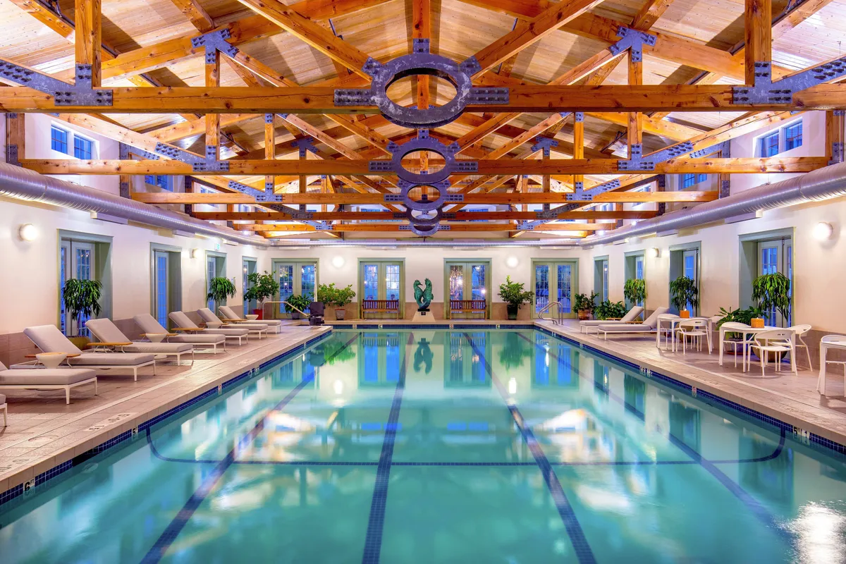 An elegant indoor swimming pool area illuminated by soft evening lights. The space features exposed wooden beams, lush decorative plants, and comfortable lounge chairs, with a nearby dining set for poolside refreshments and conversation.