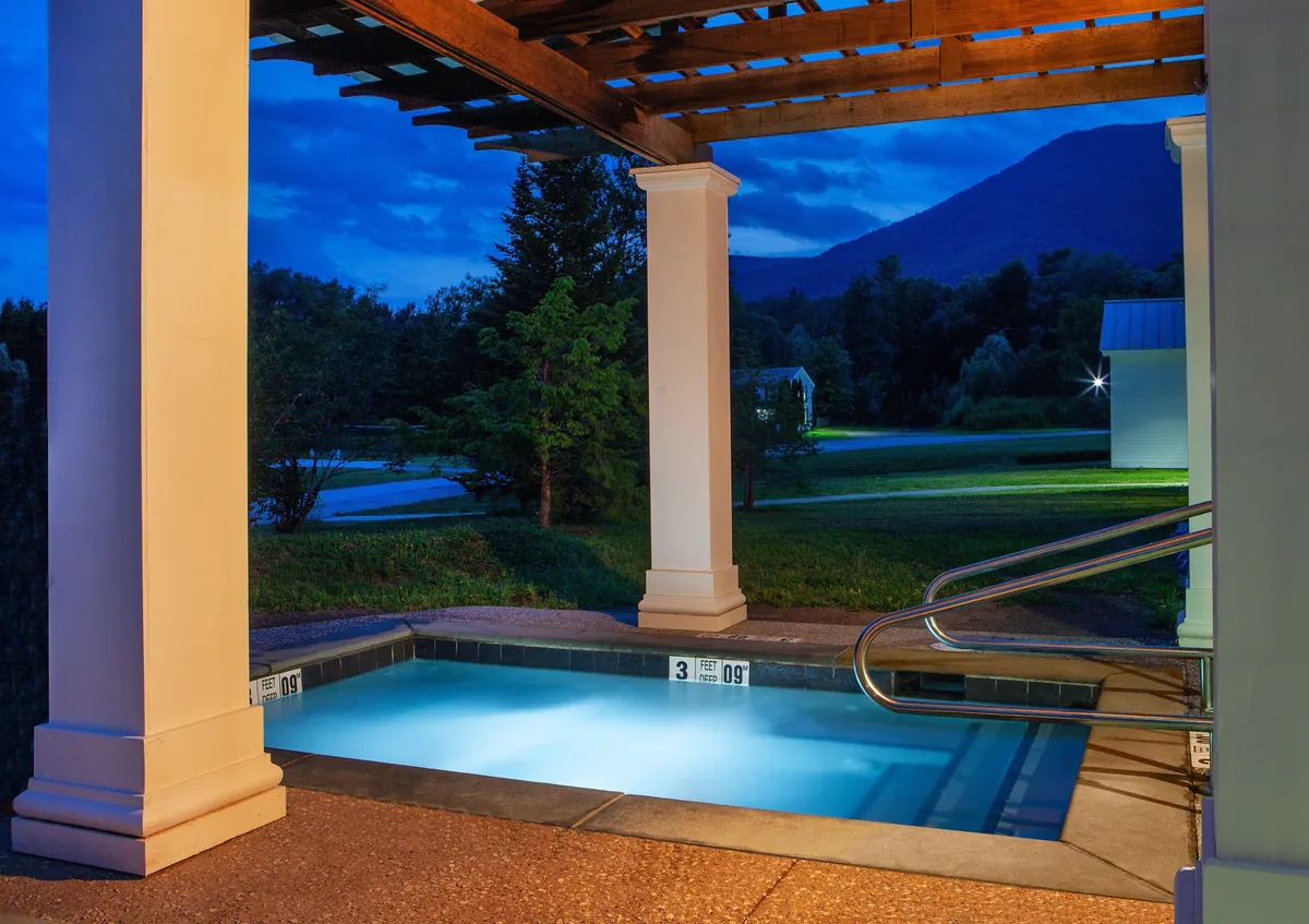 A scenic outdoor pool set against a striking mountain backdrop, with a pergola structure providing shade and atmosphere. The pool area is tranquil, showing clear water perfect for swimming or lounging. The setting suggests both daytime activity and peaceful evening relaxation, reinforcing the idea of year-round wellness opportunities.