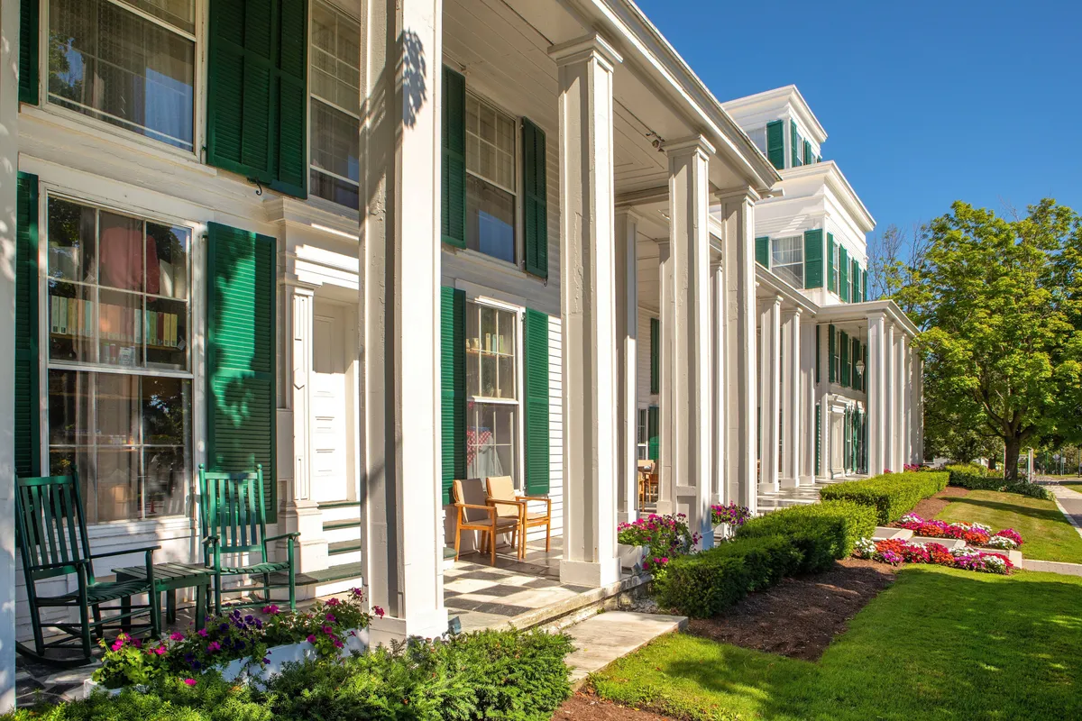 This image features the resort's exterior with classic green shutters, white columns, and rows of inviting rocking chairs on the porch. Colorful flower beds frame the entrance, enhancing the welcoming and historic vibe. The scene suggests relaxation, quiet conversation, and the timeless charm of New England architecture.