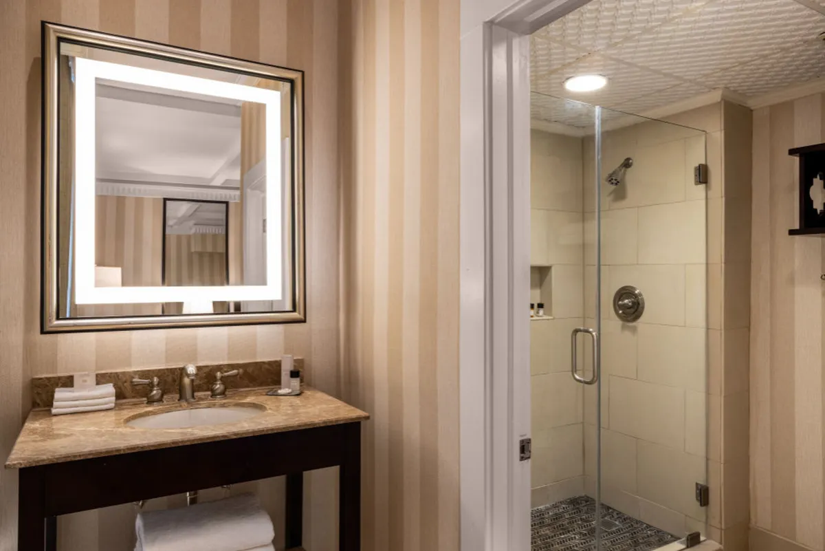 A bright, modern en-suite bathroom featuring an illuminated vanity mirror, a spacious countertop, and a walk-in shower. The space is serene, with clean lines and soft lighting that invite relaxation and rejuvenation after a long day.