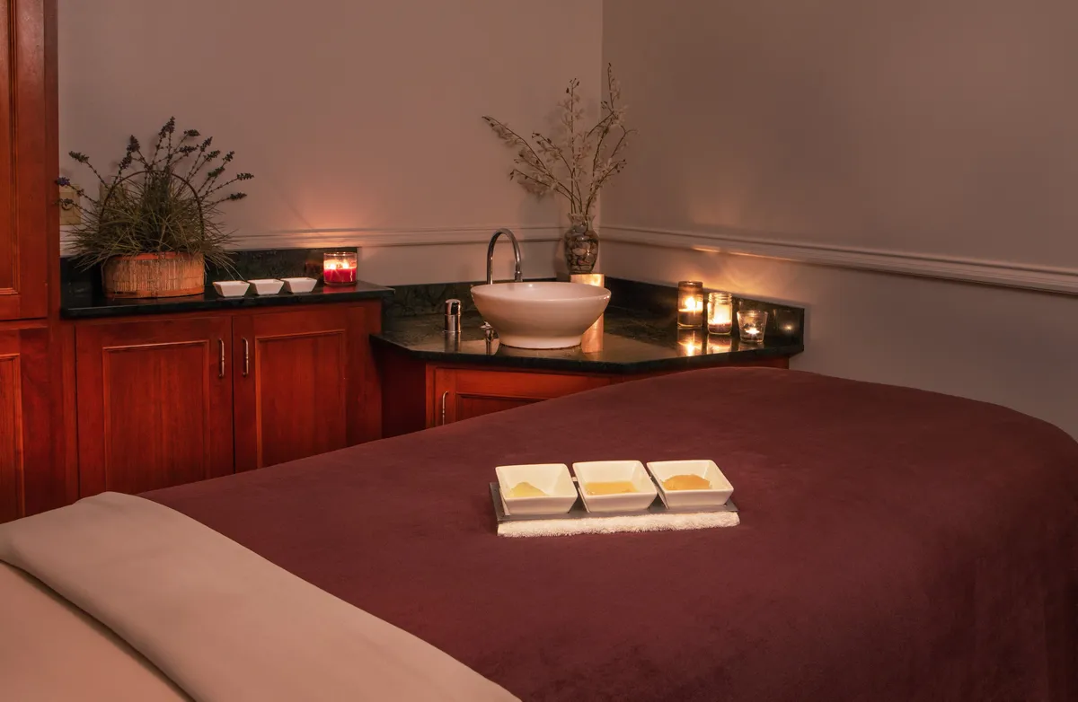 A tranquil interior spa scene featuring a massage table surrounded by decorative plants, ambient candles, a modern sink with basin, and neatly arranged spa products in bowls. The calming décor and soft lighting immediately evoke serenity and relaxation, perfectly aligning with the written emphasis on spa treatments and wellness journeys.