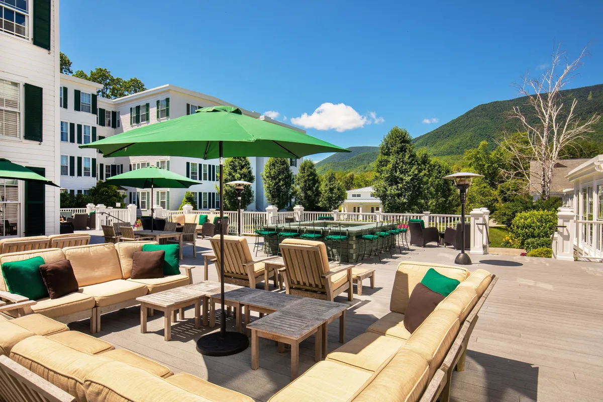 An outdoor dining area featuring green umbrellas shading tables set for groups, all positioned to take in sweeping views of the mountains. The scene is lively with guests enjoying al fresco meals and drinks, evoking the feeling of a vibrant village setting with a refreshing, open-air ambiance.