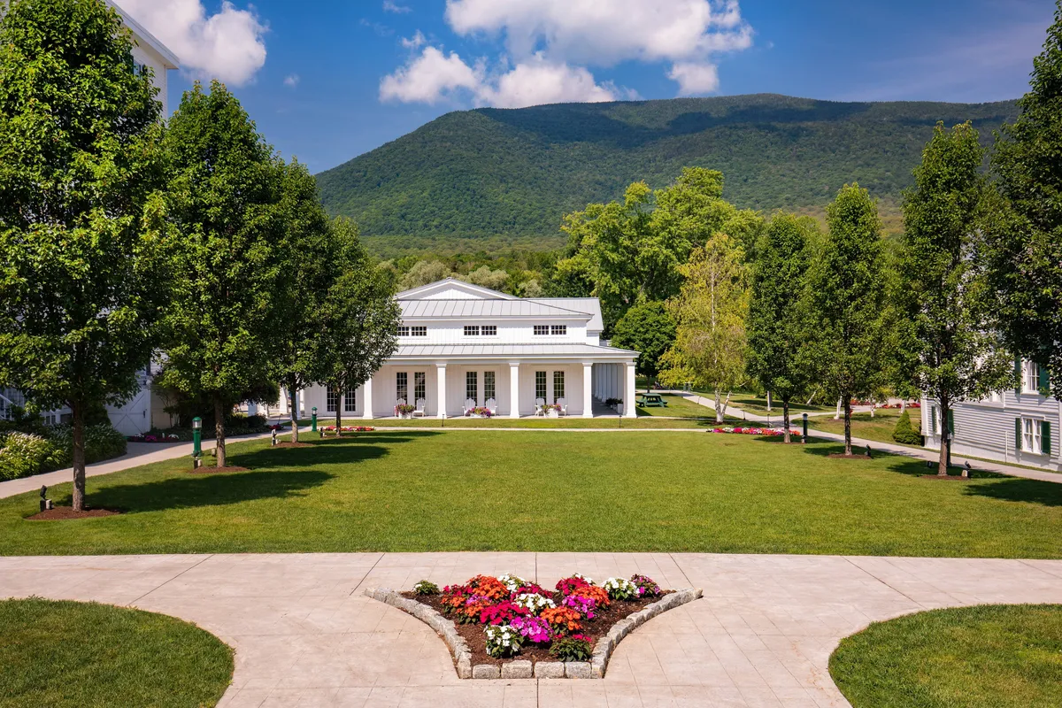 This image captures the main building of The Equinox Golf Resort & Spa with its welcoming porch, beautifully landscaped gardens, and a picturesque mountain backdrop. The scene is bathed in tranquil sunset light, suggesting both romance and relaxation. People can be seen enjoying the porch and strolling through the gardens, evoking a sense of togetherness amid natural beauty.