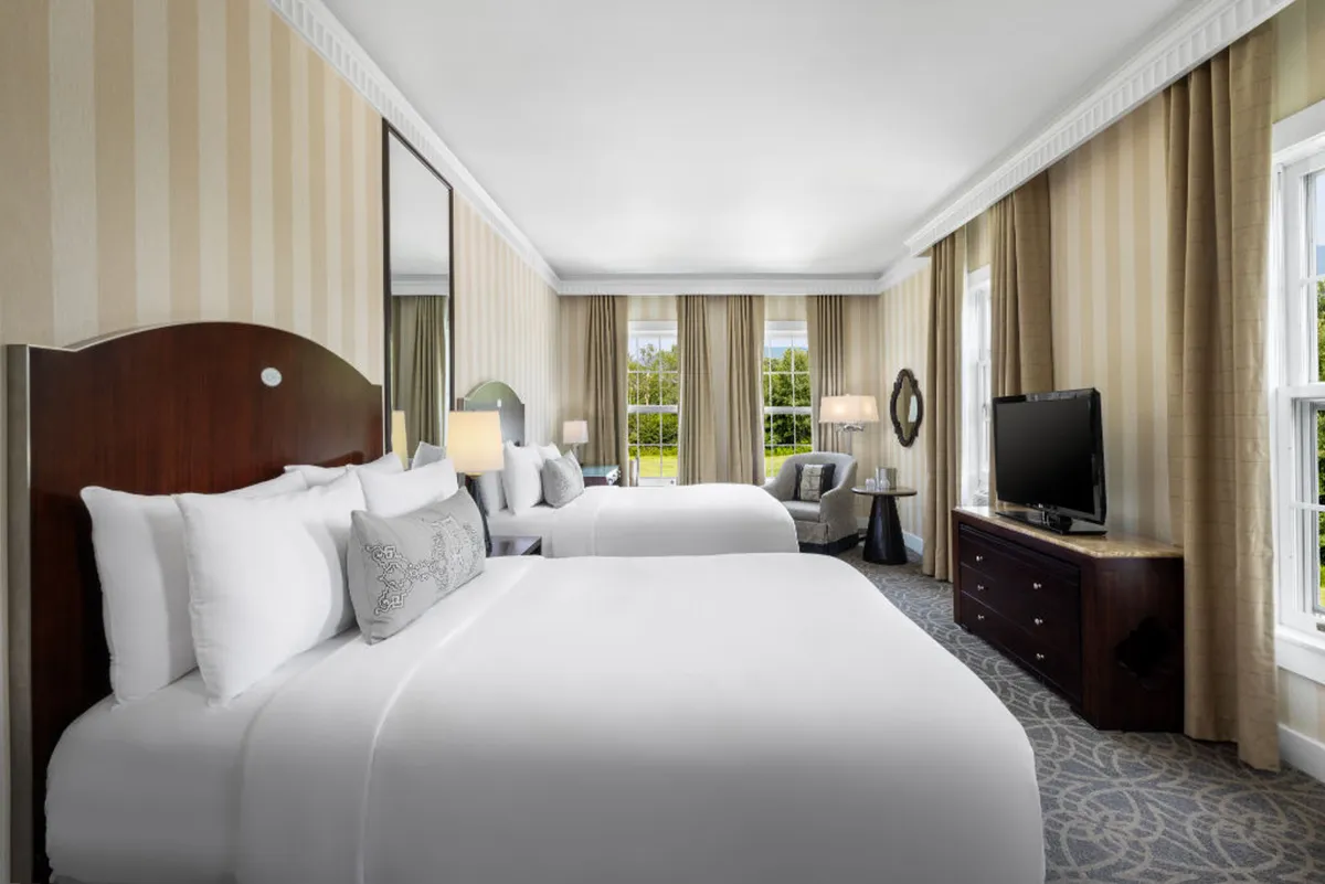 The image showcases a serene studio interior at The Equinox Golf Resort & Spa, featuring two plush beds bathed in soft natural light from expansive windows, a stylish nightstand with a glowing lamp, a decorative mirror, and a modern television. The room exudes comfort, inviting guests to relax, unwind, and soak in the tranquil ambiance.