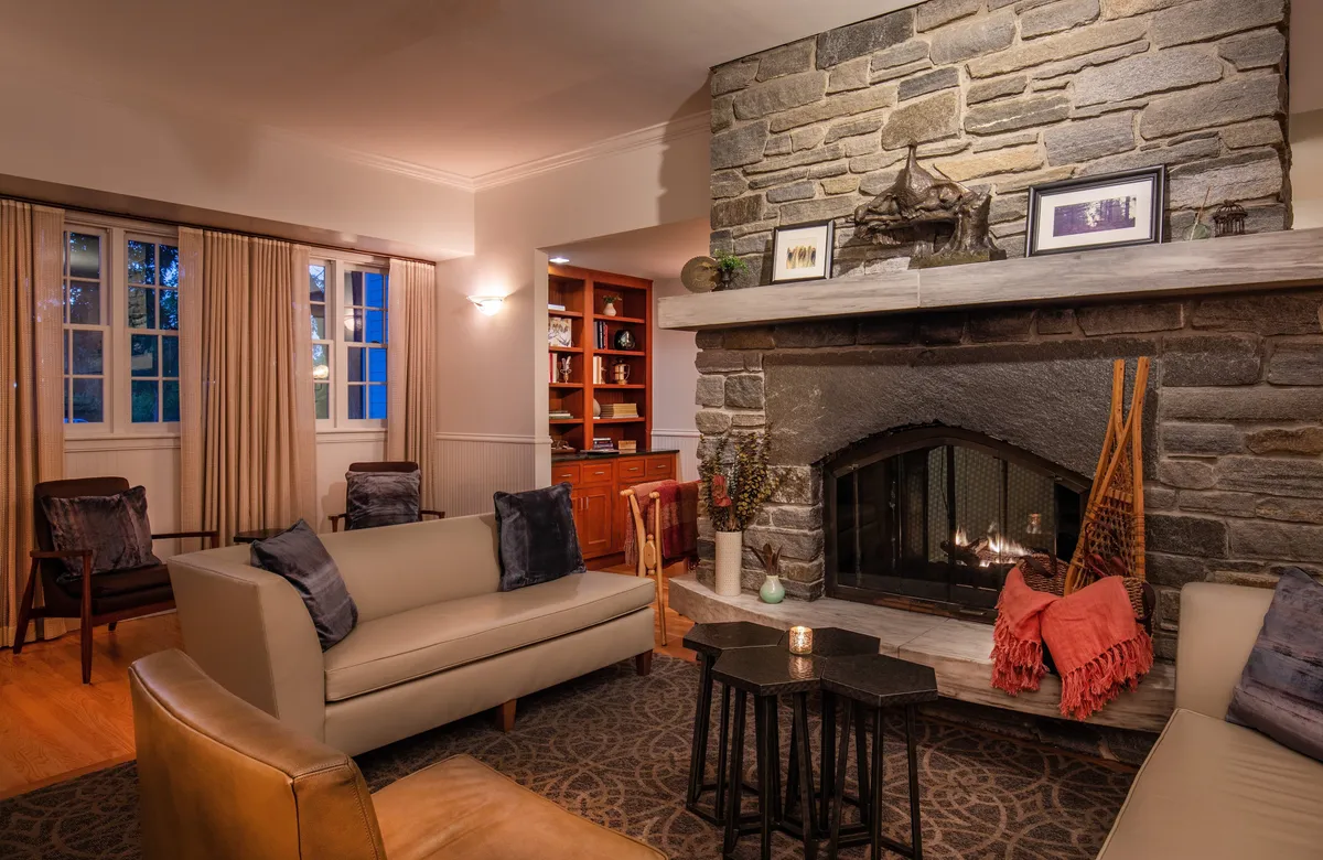 A warmly lit lounge with a striking stone fireplace as its centerpiece, surrounded by plush sofas and a bookcase filled with reading material. The inviting setting encourages guests to relax, read by the fire, or gather for intimate conversations.