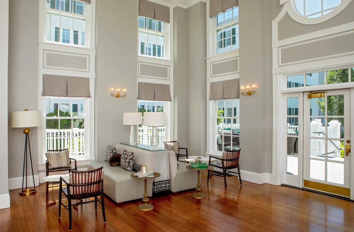 A sunlit guest room with large windows, a comfortable seating area, elegant light fixtures, and a decorative table. The space exudes a welcoming, serene mood—perfect for relaxing, reading, or enjoying refreshments in style.